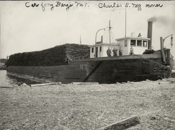 View from shoreline of a barge with four men standing on the deck. The ferry is carrying a large load.