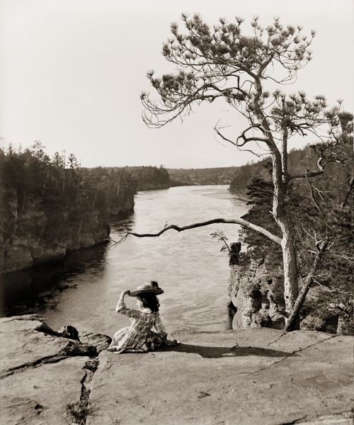 View towards High Rock from a cliff, looking down on the Wisconsin River. There is a young girl posing sitting in the foreground on the edge of the rock.