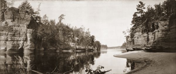 Panoramic view from shoreline of the Wisconsin River in the Wisconsin Dells. A sandy beach area is on the right, and rock formations are on both sides of the river. In the lower right hand corner are the words "JAWS OF THE WISCONSIN DELLS. Copyright 1894. by H.H. Bennett."