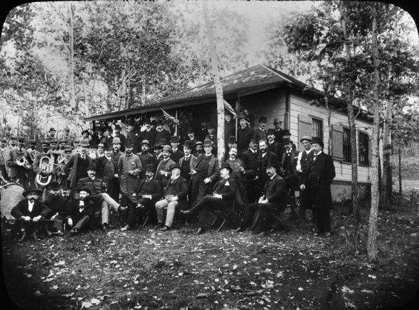 A large group of men pose outside the Rex Magnus Club. Some of the men on the left are holding musical instruments. The words "Rex Magnus" are on the roof.