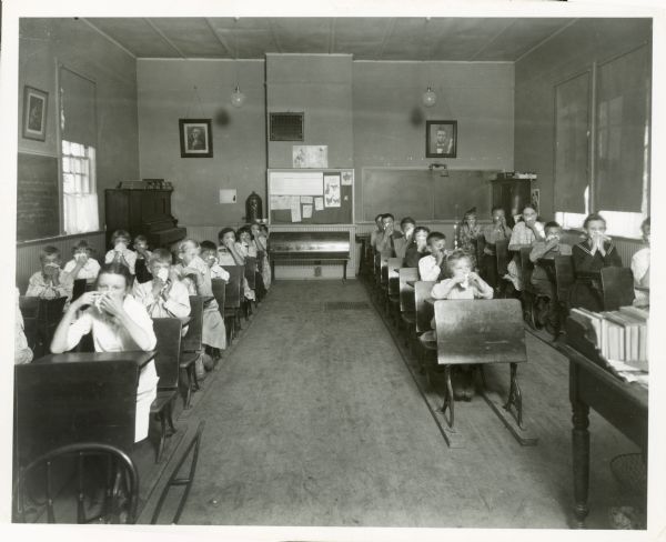 View from front of classroom of children sitting in desks at the elementary school health program, blowing their noses.