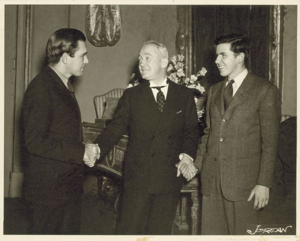 George M. Cohan is seen with two younger men. He is shaking the hand of one and holding onto the hand of the other.