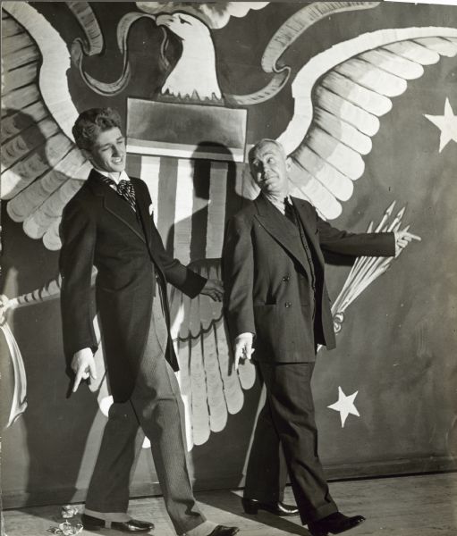 George M. Cohan and James Graham hold the same pose next to each other, with Cohen looking back toward Graham. Graham plays a younger Cohan. There is a large eagle emblem painted on the wall behind them.