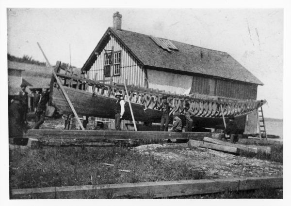 View of the steamboat <i>Mendota</i> under construction. A number of men, and one young child, are standing and posing with the boat. In the background is a building.