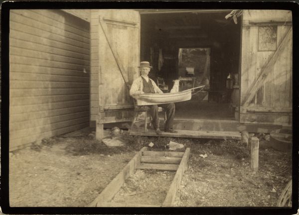Captain Paterson is seated in front of his boathouse on the 600 block of East Gorham Street. He is holding a small boat on his lap.