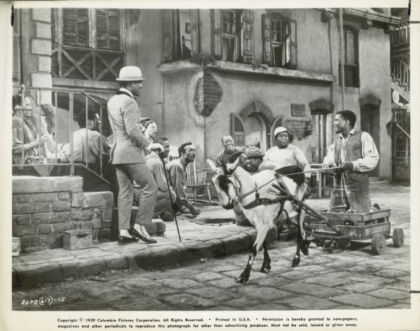 Production still from the film "Porgy and Bess." Porgy (Sidney Poitier) kneels on his cart which is pulled by a goat. He passes a group of men playing dice on the sidewalk. Sportin' Life (Sammy Davis Jr) stands to the left of the group and a number of women, including Maria (Pearl Bailey), sit on some steps.