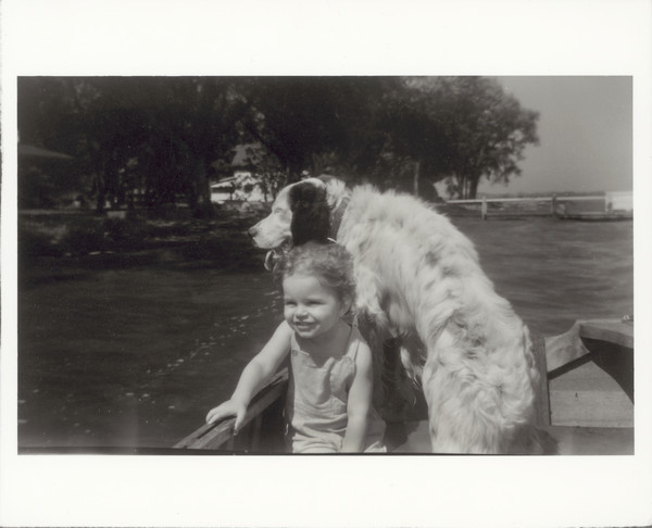 A child, probably a girl, rides in a boat with a dog. A pier and shoreline are in the background.