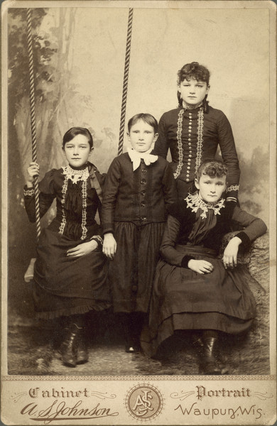 Studio portrait in front of a painted backdrop of four girls from the Sloan family. One of the girls is sitting on a swing, two are standing, and one sits on a bench.