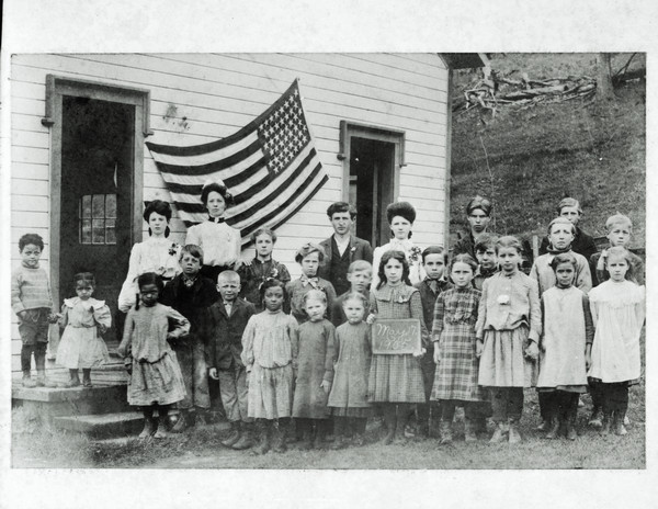 Eastman School students, along with a woman, posing outdoors in front of the school. An American flag is hanging behind them on the side of the schoolhouse. One young girl is holding a slate which has written on it: "May 17, 1905." A steep hill is behind the school on the right.