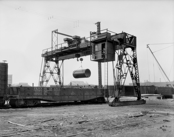 Gantry crane hoisting a large cylindrical object next to a railroad car. A man is standing in an open doorway in the operating room of the crane. The sign on the crane reads: Milwaukee Electric Crane & Mfg. Corp." In the far background is the top of a steel bridge, and industrial buildings. One building has a sign on the roof for: "Knickerbocker Masons Supply Co."