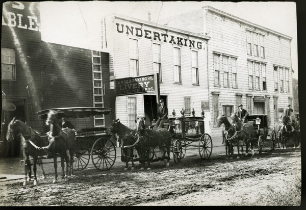 View from across unpaved street of four men with horse-drawn hearses lined up in front of an undertaking building. The other storefronts are a stable on the left, and a hardware store on the right.