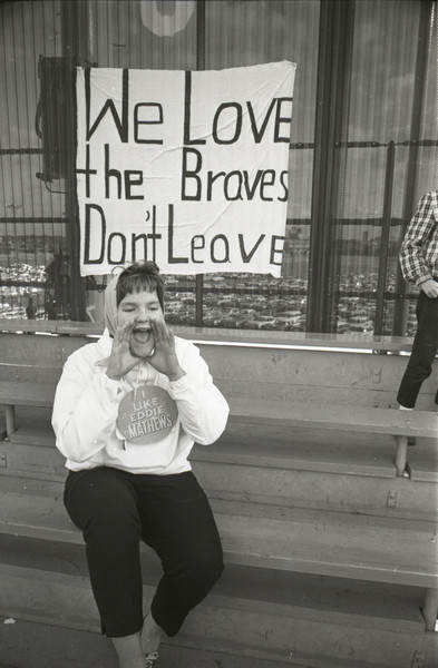 A vocal fan in the bleachers wears a sign reading "I like Eddie Mathews. She sits in front of a large sign reading "We love the Braves. Don't leave." The stadium parking lot is in the background.