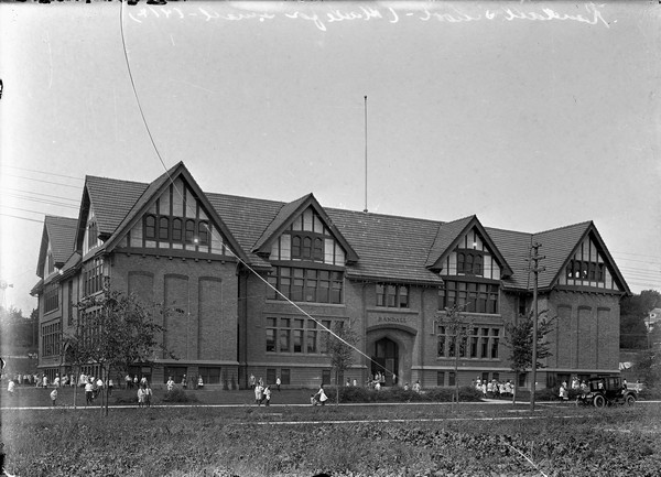 Exterior view of Randall Elementary School with students playing in the yard around the building. A car is parked along the road in front of the school. Caption reads: Randall School (Made for Small, 1914).