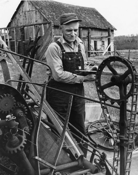 Albert Roszek, wearing overalls, posing with farm machinery. There is a barn in the background.