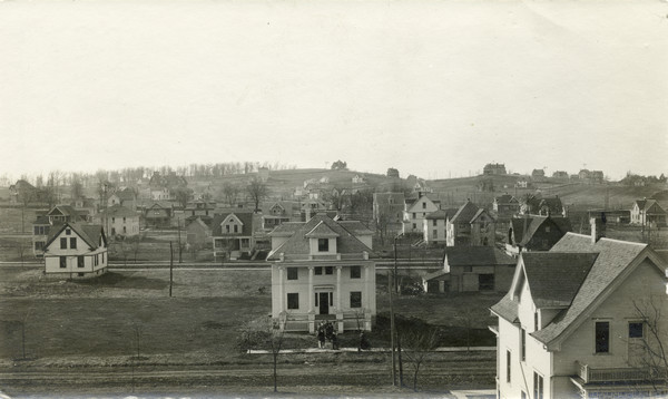 Elevated view of University Heights taken from the third floor of a home at 216 Campbell Street. Several residences can be seen in the view, including Professor Gilmore's home, 1607 Adams Street in the lower right corner. A small group of people is leaving a home,1610 Adams Street, in the center foreground. To the left is 1611 Jefferson and to the right is 1602 Jefferson.  There are windmills in the yards of some of the residences.