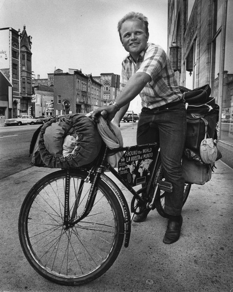 Heinz Stücke on his bicycle in front of the Milwaukee Journal building at 333 W. State Street. Stücke carries his belongings in bags attached to his bike which bears a sign with a map of the world. The names of places around the world are stenciled onto the frame and fenders of the bicycle.