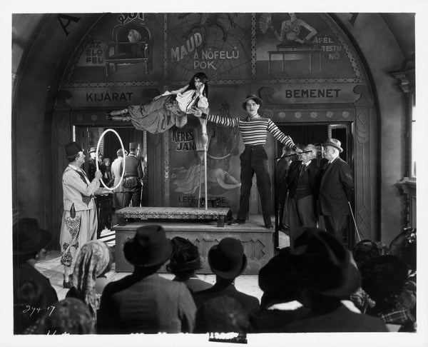 A production still from the 1927 film "The Show." Cock Robin (John Gilbert) is on a small stage with a woman floating on her side next to him. Another man stands on the floor holding a hoop.