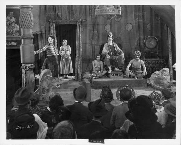 Cock Robin (John Gilbert) stands behind a big cat that is tied to a column. A man sits on a throne with a woman and man sitting on either side of him. An audience sits in front of all of them in a scene from the 1927 film "The Show."