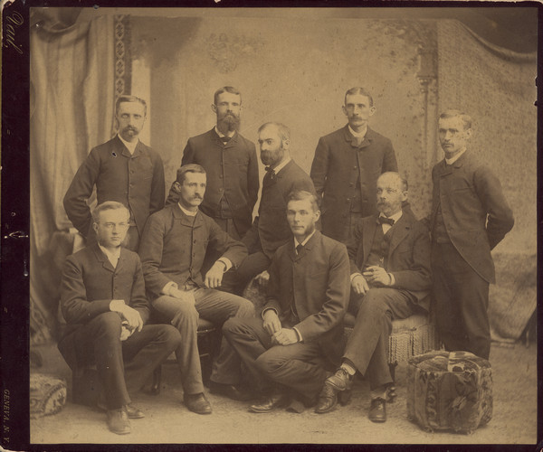Studio portrait in front of a painted backdrop of group of nine men, identified on the reverse as associates of the Geneva, New York Experiment Station. Stephen Moulton Babcock, with beard, sits in the center on the arm of a chair. Babcock was at Cornell's New York State Agricultural Experiment Station from 1881 until 1888, when he joined the Dairy Department at the University of Wisconsin. The men are wearing suits and all sport facial hair of various styles.