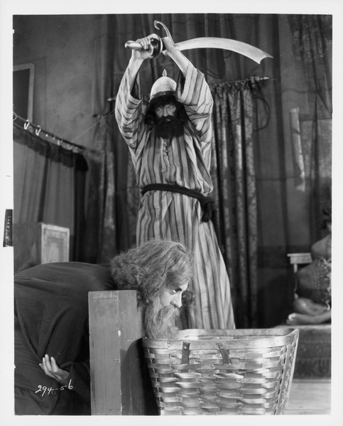 In a production still from the 1927 film "The Show," a man with a dark beard wearing a robe holds a large sword above his head. It appears that he is about to cut off the head of a man kneeling down with his head above a basket.