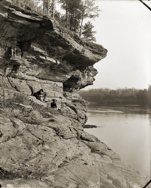 View along rockface shoreline towards a woman and two children sitting on Angel Rock. Along a cliff along the opposite shoreline are buildings and a bridge crossing the Wisconsin River.