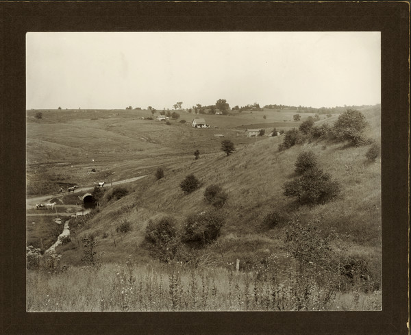 View looking down hill of open, grassy fields and houses on a hill in the distance. At the bottom of the hill in the foreground is a stream running through a stone arch culvert running under a road, and three men posing nearby with horse-drawn vehicles.