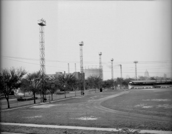 Breese Stevens Athletic Field, with lights on tall poles, and a chain link fence along the sidewalk. View looking toward E. Washington Avenue with Simon Brothers Wholesale Grocery Warehouse, 901 E. Washington Avenue, power plant chimney and storage tank in the background. The Wisconsin State Capitol is in the background on the right. (Section of panorama.)