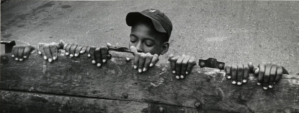 Slightly elevated view of a boy in a baseball cap peeking over a fence. There are several sets of hands curling over the top of the fence on either side of the boy.