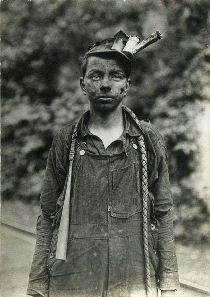 Boy dressed in work clothes and covered with soot from working in a mine. He wears a hat with a rudimentary lantern on top.