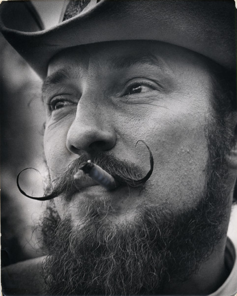 Allen De Karske shows off his full beard and waxed, handlebar moustache. De Karske is also wearing a hat and is smoking a cigarette.