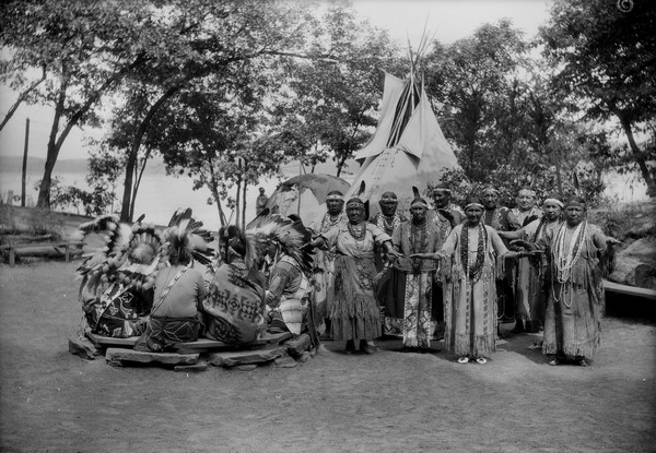 Ho-Chunk women perform the Swan Dance. There is a group of men sitting in a circle next to them.