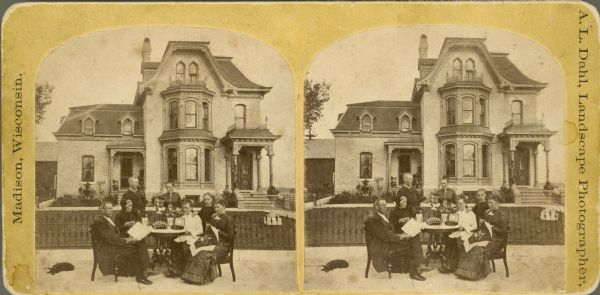 Martin K. Dahl, far left, sits with three women around a table which has been set in front of an iron fence which encloses a manicured yard. Two of the women are doing needlework. Another man, woman and child stand behind the others. The standing woman holds a pitcher and glass.  A dog rests on the ground to the left of the group. The two and one-half story brick Second Empire style brick house has a two-story bay and two porches which are topped by decorative ironwork. A barn is  behind and to the left of the house.