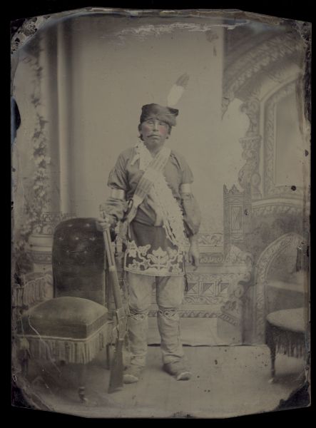 Full-length studio portrait in front of a painted background of Little Soldier. He is standing next to a chair and holding a firearm. Hand-coloring on cheeks. Notes on back of tintype/ferrotype read: "Fur turban; Eagle feather erect; Woodland floral breech cloth beaded; geometric garters beaded; German silver arm bands; bugle & shell necklace; Winnebago moccasins; Buckskin breeches fringed at sides."