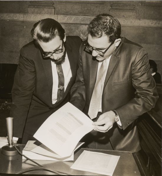 University of California molecular biologist, Dr. Robert Riseborough, left, confers during a break in the DDT hearings with Victor J. Yannacone, the attorney representing conservation groups who filed a petition to ban the use of DDT in Wisconsin.
A decorative architectural border can be seen on the wall behind the men.