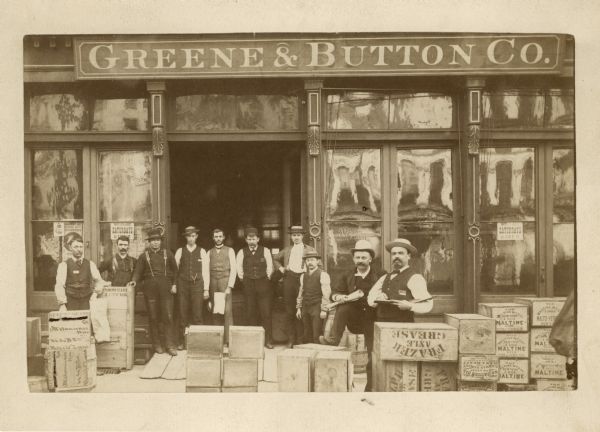 A group of men, possibly employees, pose in front of Greene & Button Co. store among boxes and crates of various goods.