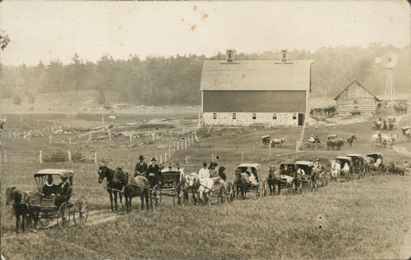 View of a procession of horse-drawn carriages along a dirt road through a field. The second carriage from the left is a hearse. There is a farm in the background.