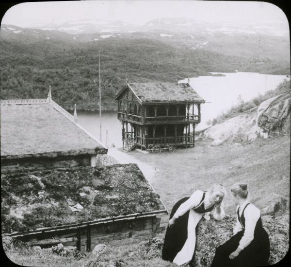 View of a mountaintop house and other buildings with sod roofs. Two women, who are possibly Swiss or Norwegian, are in the foreground sitting on a steep hill. Below are the buildings, which are high above a river. In the far background is the far shoreline with tree-covered hills rising to snow-capped mountains.