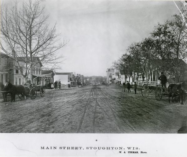 View down Main Street. On the left a man is driving a wagon with a team of two horses, and on the right a man stands on a wagon pulled by one horse. Houses and storefronts are along both sides of the street.