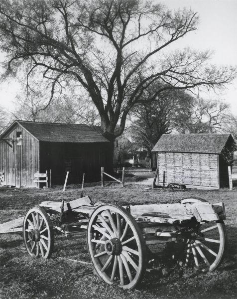 The frame of a broken wagon sits in a field. In the background are in several farm buildings and a farmhouse.