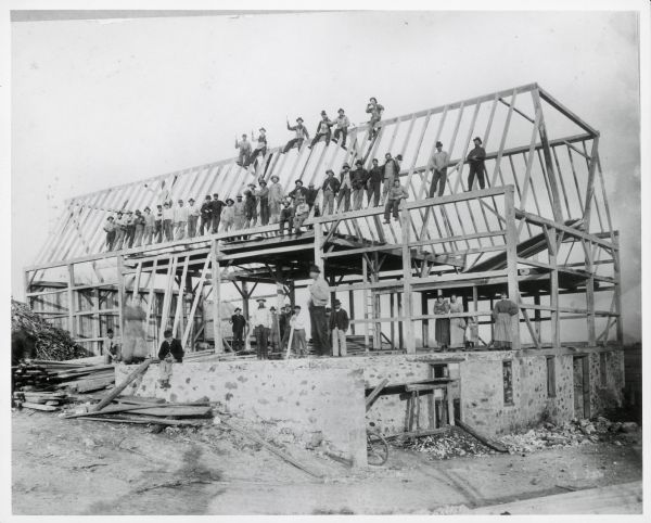 A large group of men pose on the wooden framework of a barn they're building on Holy Hill Road, which has a stone foundation. On the far right three women and a young girl stand on top of the stone foundation of the barn.