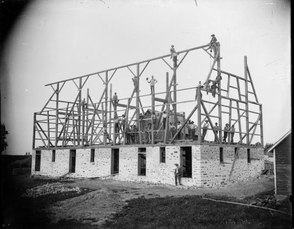 A large group of men pose on the wooden framework of a barn that they are building.