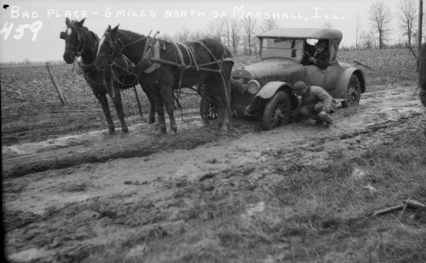An automobile is stuck in the mud on a rural road. The driver leans out of the window to observe a man putting material for traction under the front, left wheel. Another man can be seen hitching the two harnessed horses to the front of the car, which is covered with mud. The driver is wearing a suit and hat, the other two men are wearing work clothes. In the background is a harvested corn field behind a fence with trees in the distance. The horses stand patiently, waiting to pull the car our of the mire. Text at the top of the image reads, "Bad Place - 6 miles north of Marshall, Ill."