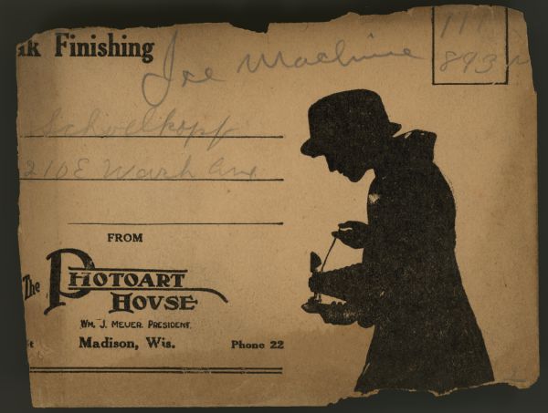 The tattered remains of a processing envelope from The Photoart House. Handwritten on the envelope is "Ice Machine, 111, 893," "Schoelkopf, 210 E Wash Ave." Above is printed "[Xxx]ak Finishing," below is "From the Photoart House, Wm. J. Meuer. President. Madison Wis. Phone 22." On the right is a waist-up silhouette of a woman photographer wearing a coat and hat, holding a camera with bellows extended, release cord in her hand.