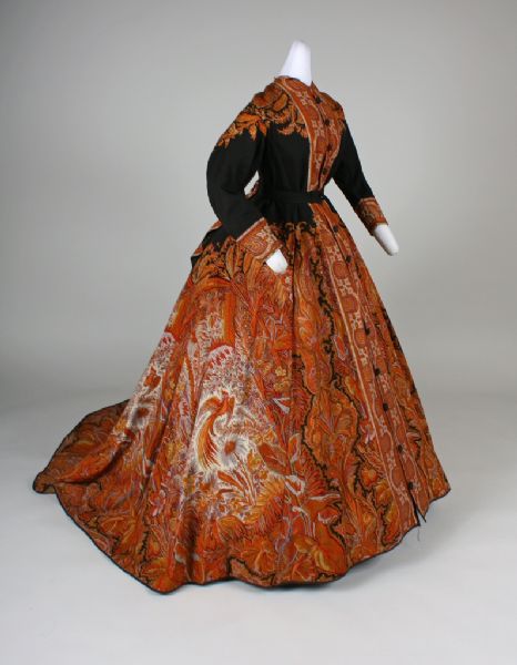 Three-quarter view of a lightweight wool dress cut from a shawl, in cream, rust and indigo paisley with a black center. The paisley has a bird-of-paradise design. It has covered buttons on the front down to the floor and is made to be worn with a hoop. The dress is displayed on a mannequin.