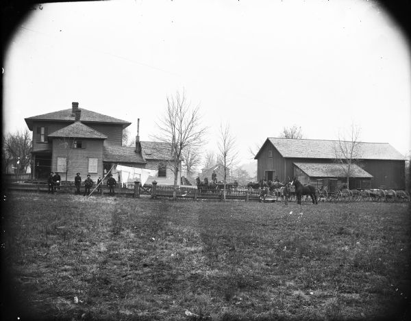 View across grassy field towards a large group of men, horses and horse-drawn vehicles posing along a wooden fence with a gate. There is a farmyard with a farmhouse and farm buildings behind them. One man is standing on the porch. Laundry is hanging on a line strung between two trees near the fence.