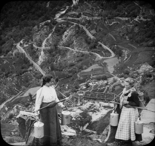 Two Norwegians milkmaids stand together on Grjotlid Road, holding cans of milk on yokes around their necks. They are standing at the edge of a steep hill, with a valley far down below them.