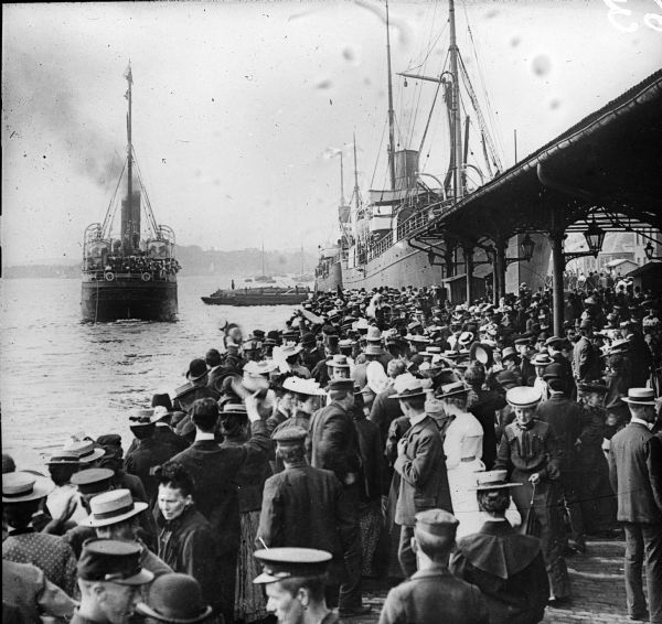 Elevated view of a crowd of Norwegian people standing at a dock looking out at the ships.