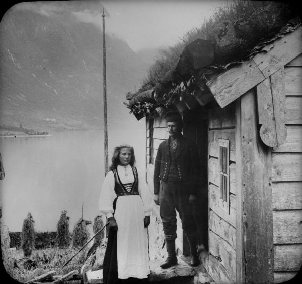 A farming couple pose together on the steps of a building with a sod roof in Nordfjord country. In the background is a lake and a mountain on the far shoreline.