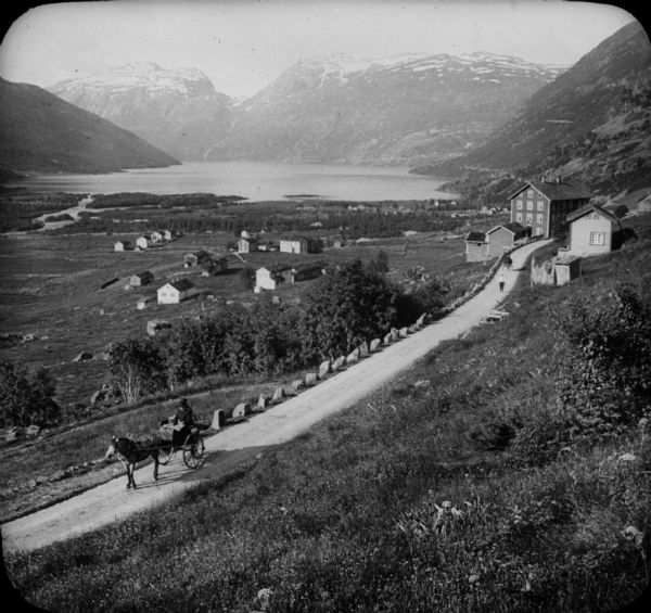 Elevated view of a town and body of water in a valley in Roldal, Norway. Two people in a horse-drawn vehicle are in the foreground on the road leading from the town.