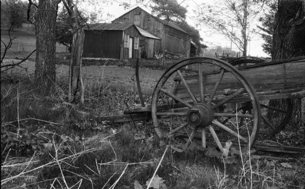 A view of the William Ruggles farm with an old farm wagon in the foreground. A number of farm buildings are in the background, with a fence and hill on the left. Telephone poles and an agricultural implement are in the center background.
